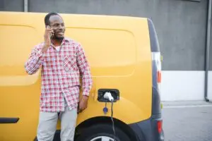 African American man stands next to an electric delivery van by an EV charging station.