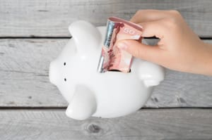 A boy's hand inserting a fifty-dollar banknote into a piggy bank.